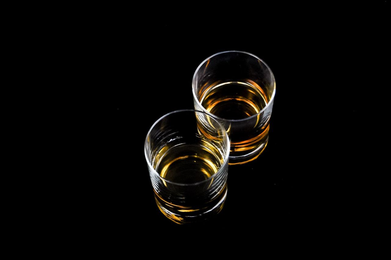 our-story Two whiskey glasses with amber liquid reflecting on a glossy black surface.