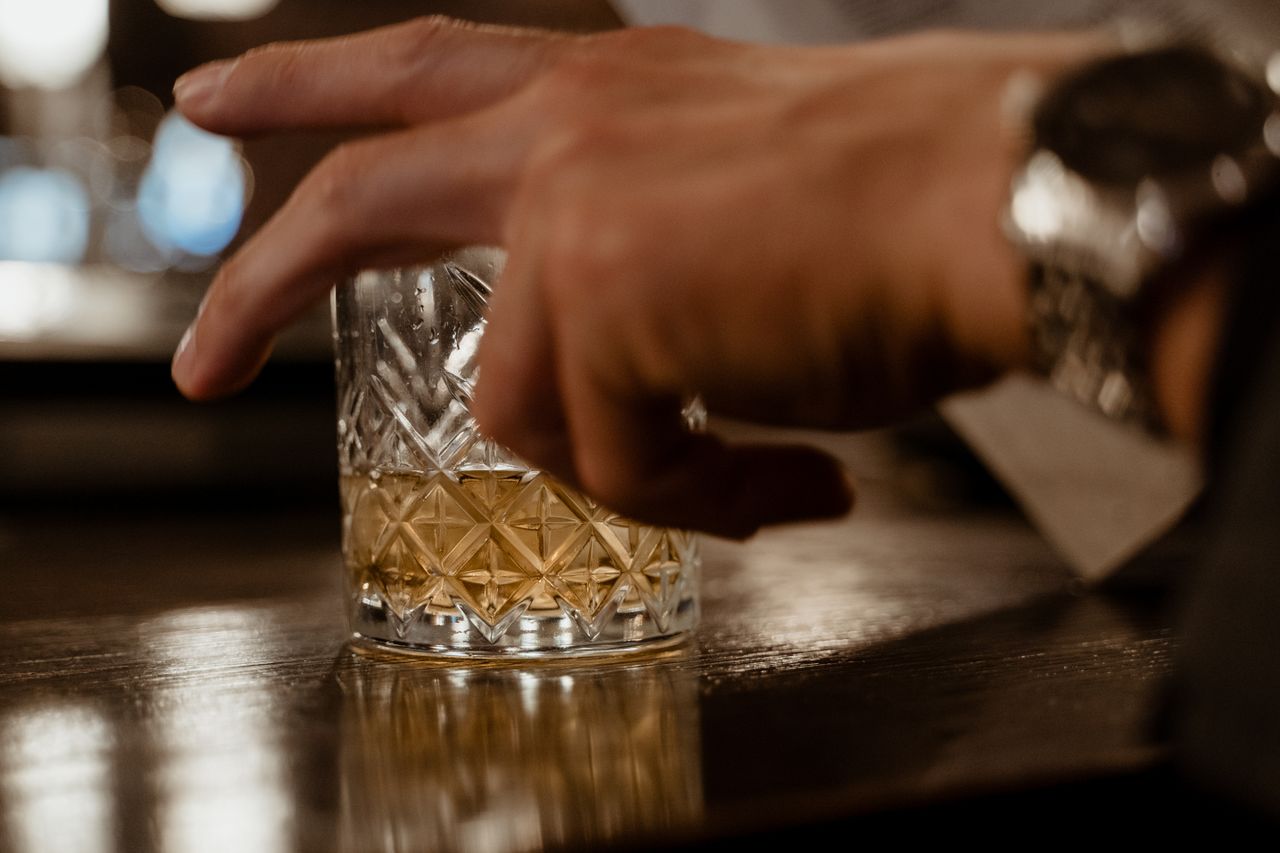 who-we-are Close-up of a hand near a crystal glass of whisky on a bar counter, exuding elegance and style.
